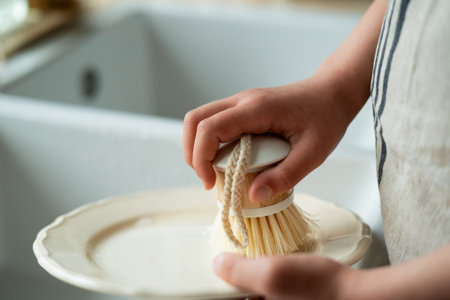 Closeup of children's hands washing a plate near the sink in the kitchen. Little girl holding an ecofriendly dishwashing brush.の写真素材