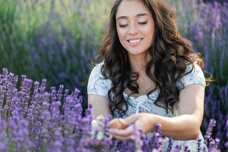 Young beautiful woman with long dark hair in a hat picking lavender flowers in a field. Holds a jar of essential oil in her hands.の写真素材