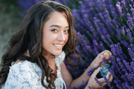 Young beautiful woman with long dark hair in a hat picking lavender flowers in a field. Holds a jar...