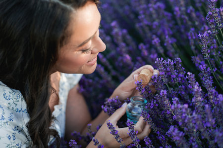 Young beautiful woman with long dark hair in a hat picking lavender flowers in a field. Holds a jar...