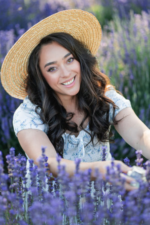 Young beautiful woman with long dark hair in a hat picking lavender flowers in a field. Holds a jar of essential oil in her hands.の写真素材