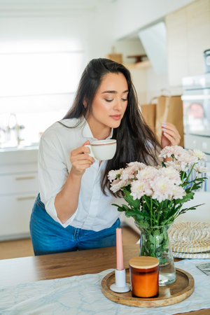 Young woman with brown hair drinking hot coffee at home in a bright kitchen.の写真素材