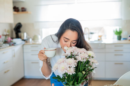 Young woman with brown hair drinking hot coffee at home in a bright kitchen.の写真素材