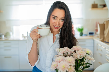Young woman with brown hair drinking hot coffee at home in a bright kitchen.の写真素材