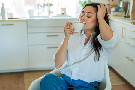 Young woman with brown hair drinking hot coffee at home in a bright kitchen.の写真素材