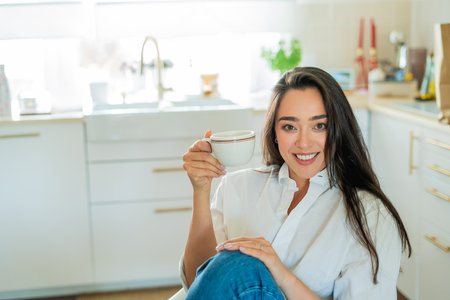Young woman with brown hair drinking hot coffee at home in a bright kitchen.の写真素材