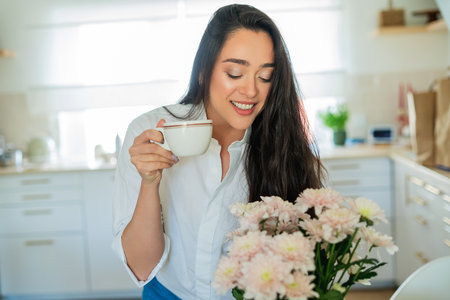 Young woman with brown hair drinking hot coffee at home in a bright kitchen.の写真素材