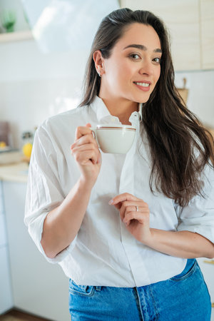 Young woman with brown hair drinking hot coffee at home in a bright kitchen.の写真素材