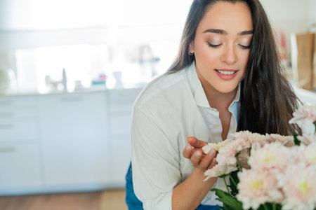 Young woman with dark hair puts a bouquet of pink flowers in a vase at home in a bright kitchen. Cozy.の写真素材