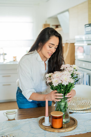 Young woman with dark hair puts a bouquet of pink flowers in a vase at home in a bright kitchen. Cozy.の写真素材