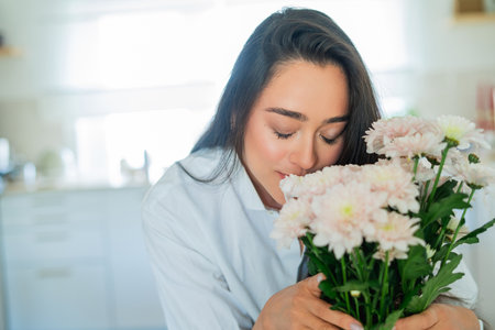 Young woman with dark hair puts a bouquet of pink flowers in a vase at home in a bright kitchen. Cozy.の写真素材