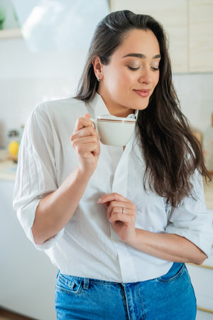 Young woman with brown hair drinking hot coffee at home in a bright kitchen.の写真素材