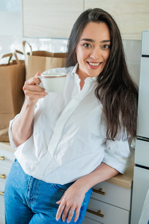Young woman with brown hair drinking hot coffee at home in a bright kitchen.の写真素材