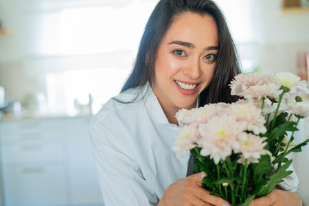 Young woman with dark hair puts a bouquet of pink flowers in a vase at home in a bright kitchen. Cozy.の写真素材