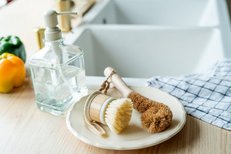 Plate with an ecobrush for washing dishes near the sink, detergent in a glass bottle, a towel. Bright kitchen interior.の写真素材