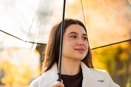 Rainy season. Young woman in cozy coat with umbrella in autumn park.の写真素材