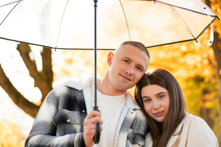 Rainy season. Young beautiful couple in love walking under an umbrella in an autumn park. Yellow leaves of trees.の写真素材