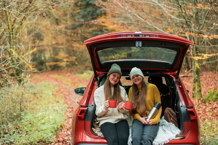 Teenage sisters drinking hot tea from a large thermos in the trunk of a red car in an autumn forest....