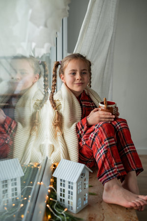 Little girl in pajamas sitting by the window drinking hot cocoa from a Christmas cup with a gingerbread man. Holidaysの写真素材