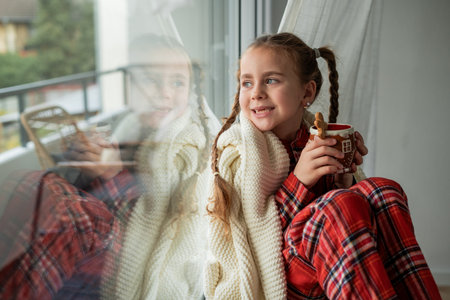 Little girl in pajamas sitting by the window drinking hot cocoa from a Christmas cup with a gingerbread man. Holidaysの写真素材