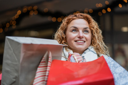 Red haired happy woman holding a red bag in her hands, shopping for Christmas gifts at a European holiday market.の写真素材