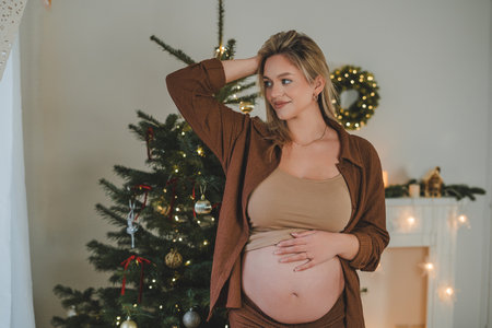 Young pregnant woman in pajamas hugging her belly near the Christmas tree. Waiting for the baby. Motherhood.の写真素材