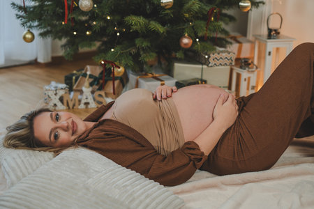 Young pregnant woman in pajamas hugging her belly near the Christmas tree. Waiting for the baby. Motherhood.の写真素材