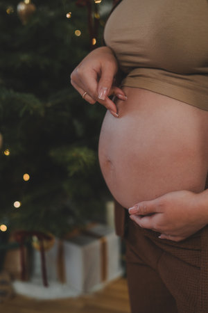 Young pregnant woman in pajamas hugging her belly near the Christmas tree. Waiting for the baby. Motherhood.の写真素材