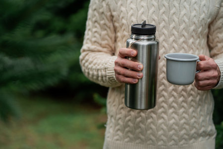 Young man with beard drinks hot tea from thermos at an outdoor setting. Winter.