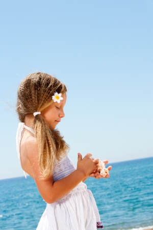 Cute girl in white dress holding shells on sunny beach の写真素材