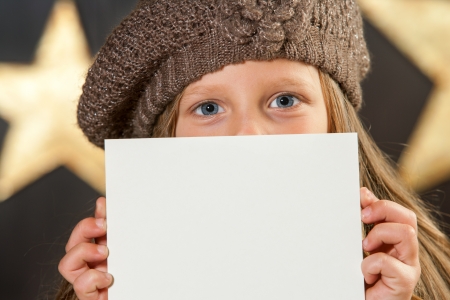 Cute girl with beanie showing big blue eyes behind blank card の写真素材