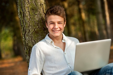 Portrait of handsome teenager sitting against tree with laptop in park.の写真素材