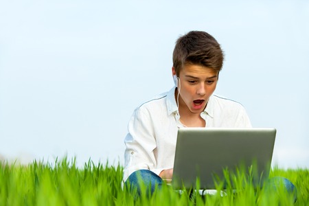 Surprised teen sitting with laptop in green grass field.の写真素材