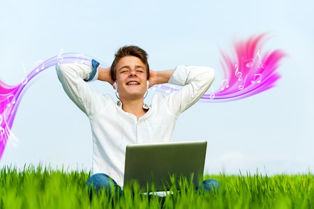 Conceptual portrait of teen boy in green grass field listening to music on laptop.の写真素材