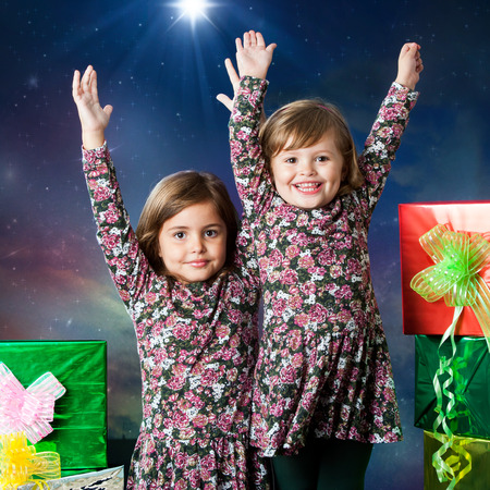 Close up portrait of two happy young sisters raising hands next to gifts. Studio portrait with winter background.の写真素材