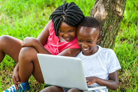 Close up portrait of happy African boy and girl playing on laptop under tree.の写真素材