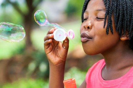 Close up portrait of cute African girl with braids blowing bubbles in park.の写真素材