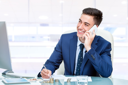 Close up portrait of Attractive young accountant talking on smart phone in office. Young man at desk in front of computer filling out financial documents.の写真素材