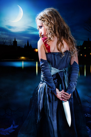 Close up portrait mysterious gothic girl holding knife behind back. Girl standing with big knife behind back at dusk. Medieval cityscape in background with reflection on dark water.の写真素材
