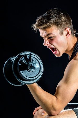 Close up action portrait of teen boy working out with dumbbell. Handsome young man isolated against black background.の写真素材