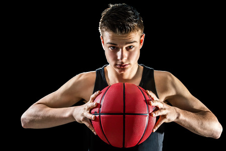 Close up studio portrait of handsome young basketball player holding inflated orange ball.Isolated against black background.の写真素材