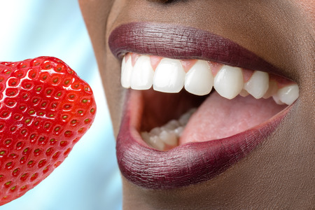 Extreme close up of african female mouth showing perfect teeth.Appetizing red strawberry  next to...