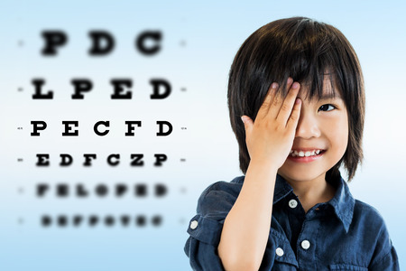 Close up portrait of cute little asian boy doing eye test.Kid closing one eye with hand against alphabetical out of focus test chart in background.