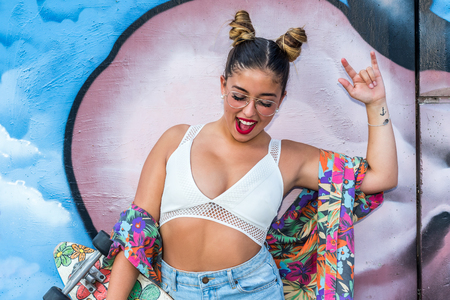 Close up portrait of cute pony tailed girl teen skateboarder doing love hand sign Against colorful graffiti wall.の写真素材