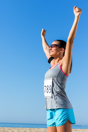 Close up portrait of young female athlete with start number showing winning attitude.Girl raising arms at sea front.の写真素材
