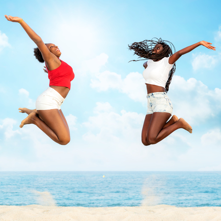 Full  length action portrait of two african girlfriends jumping together on beach. Girls screaming with hands in air.の写真素材