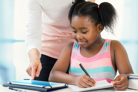 Close up portrait of little african girl struggling with school work.Caucasian teacher showing results on digital tablet at desk.の写真素材