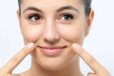 Extreme close up portrait of cute young woman pointing at wrinkles below cheek. Girl with charming smile looking aside.Isolated on white background.の写真素材
