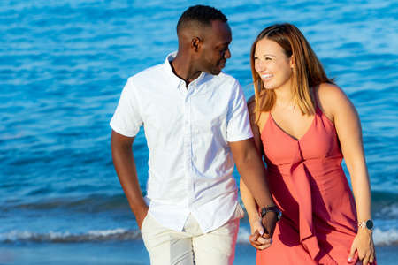 Close up portrait of young diverse couple in casual wear holding hands on beach. African man and caucasian girl against blue sea background.の写真素材