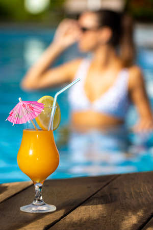 Close up of appetizing fruit cocktail on wooden deck. Out of focus background of woman in swimming...